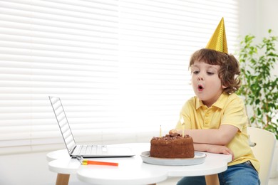 Photo of Birthday celebration. Little boy blowing candles on cake while having video call via laptop at table indoors. Space for text