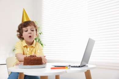 Photo of Birthday celebration. Little boy blowing candles on cake while having video call via laptop at table indoors. Space for text