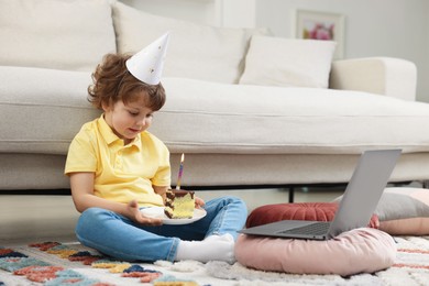 Photo of Birthday celebration. Little boy with piece of cake having video call via laptop at home