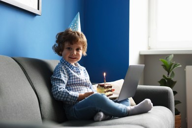 Photo of Birthday celebration. Little boy with piece of cake having video call via laptop on sofa at home