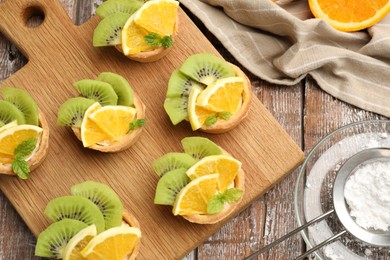 Photo of Tartlets with fruits, mint and powdered sugar on wooden table, flat lay. Delicious dessert