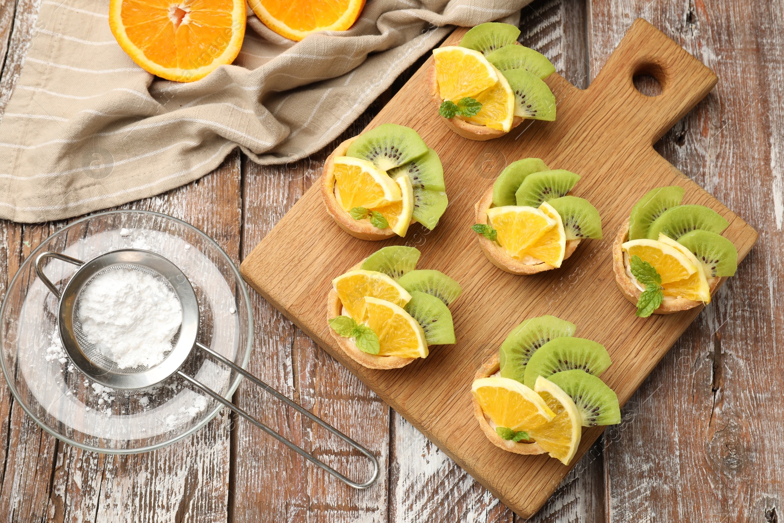 Tartlets with fruits, mint and powdered sugar on wooden table, flat lay. Delicious dessert Photo of Tartlets with fruits, mint and powdered sugar on wooden table, flat lay. Delicious dessert