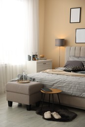 Photo of Comfortable bed with soft linens, slippers, pouf and side table in room. Home textile