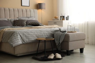 Photo of Comfortable bed with soft linens, slippers, pouf and side table in room. Home textile