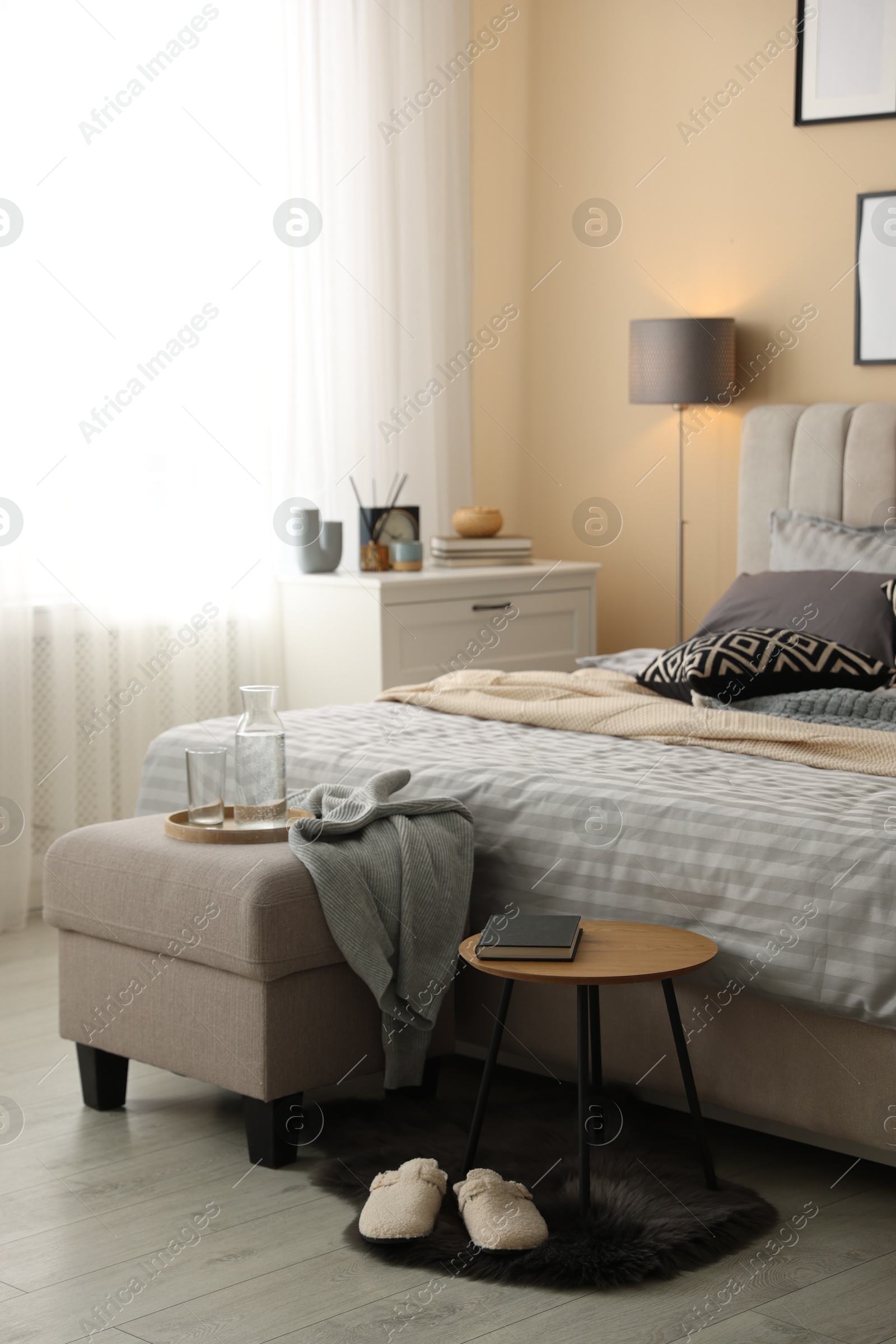 Comfortable bed with soft linens, slippers, pouf and side table in room. Home textile Photo of Comfortable bed with soft linens, slippers, pouf and side table in room. Home textile