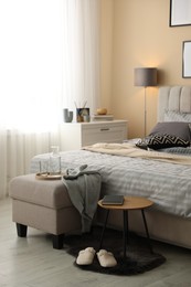 Photo of Comfortable bed with soft linens, slippers, pouf and side table in room. Home textile