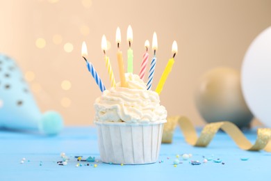 Photo of Burning candles in cupcake, sprinkles and party decor on light blue table against beige background with blurred lights