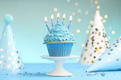 Photo of Burning candles in cupcake, sprinkles and party hats on light blue table against turquoise background with blurred lights
