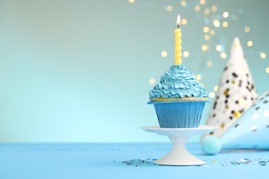 Photo of Burning candle in cupcake, sprinkles and party hats on light blue table against turquoise background with blurred lights, space for text
