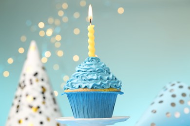 Photo of Burning candle in cupcake on cake stand and party hats against light blue background with blurred lights