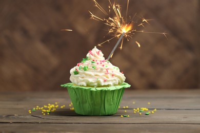 Photo of Burning sparkler in cupcake and sprinkles on wooden table