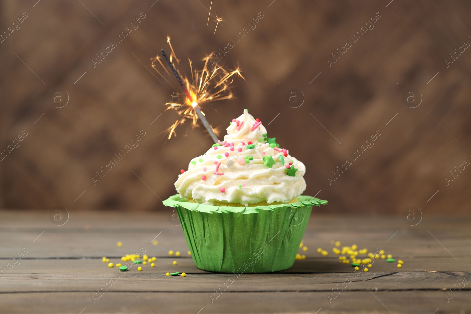 Photo of Burning sparkler in cupcake and sprinkles on wooden table
