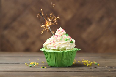 Photo of Burning sparkler in cupcake and sprinkles on wooden table
