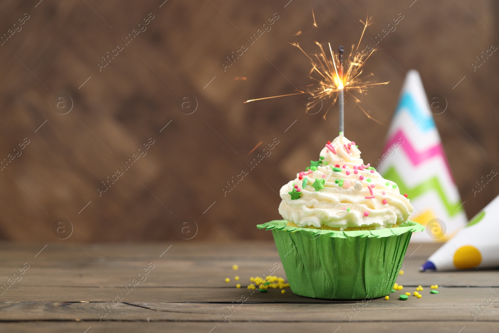 Photo of Burning sparkler in cupcake, sprinkles and party hats on wooden table, space for text