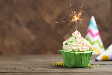 Photo of Burning sparkler in cupcake, sprinkles and party hats on wooden table, space for text