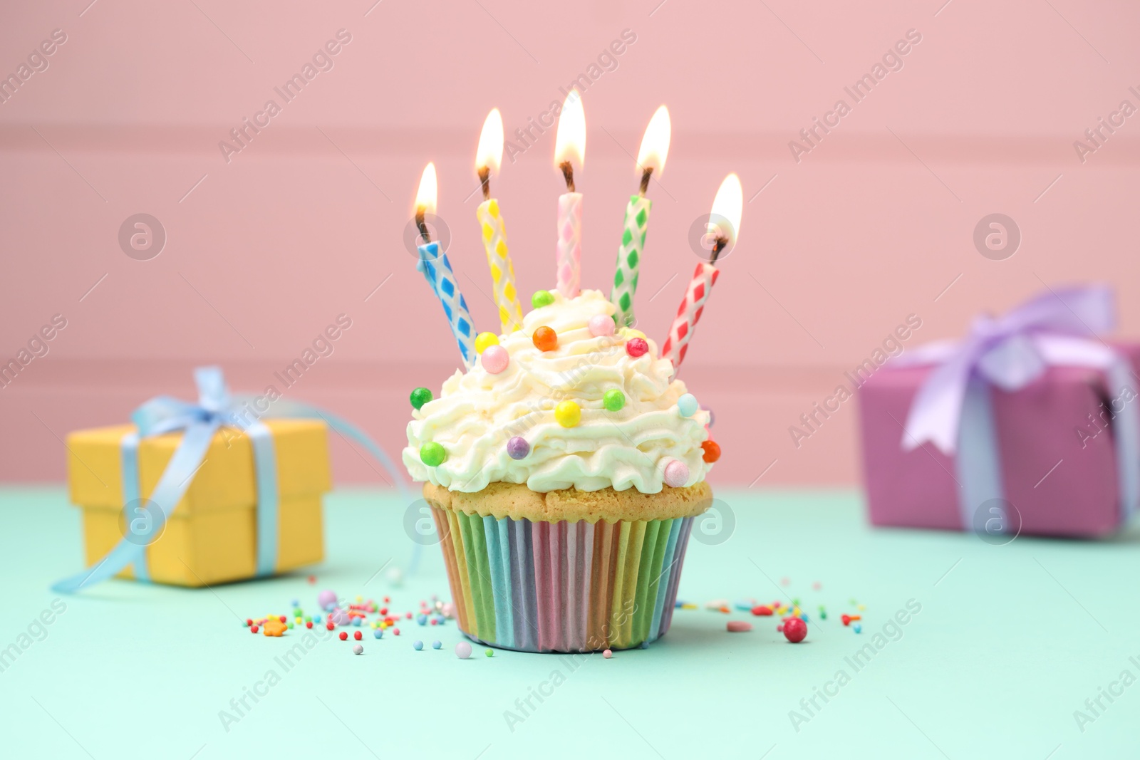 Photo of Burning candles in cupcake, sprinkles and gift boxes on turquoise table against pink background