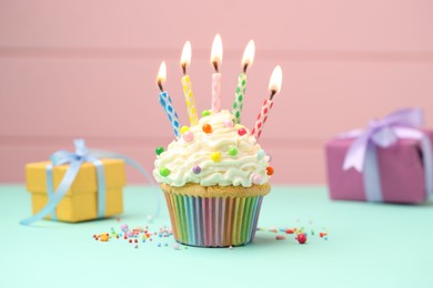 Photo of Burning candles in cupcake, sprinkles and gift boxes on turquoise table against pink background