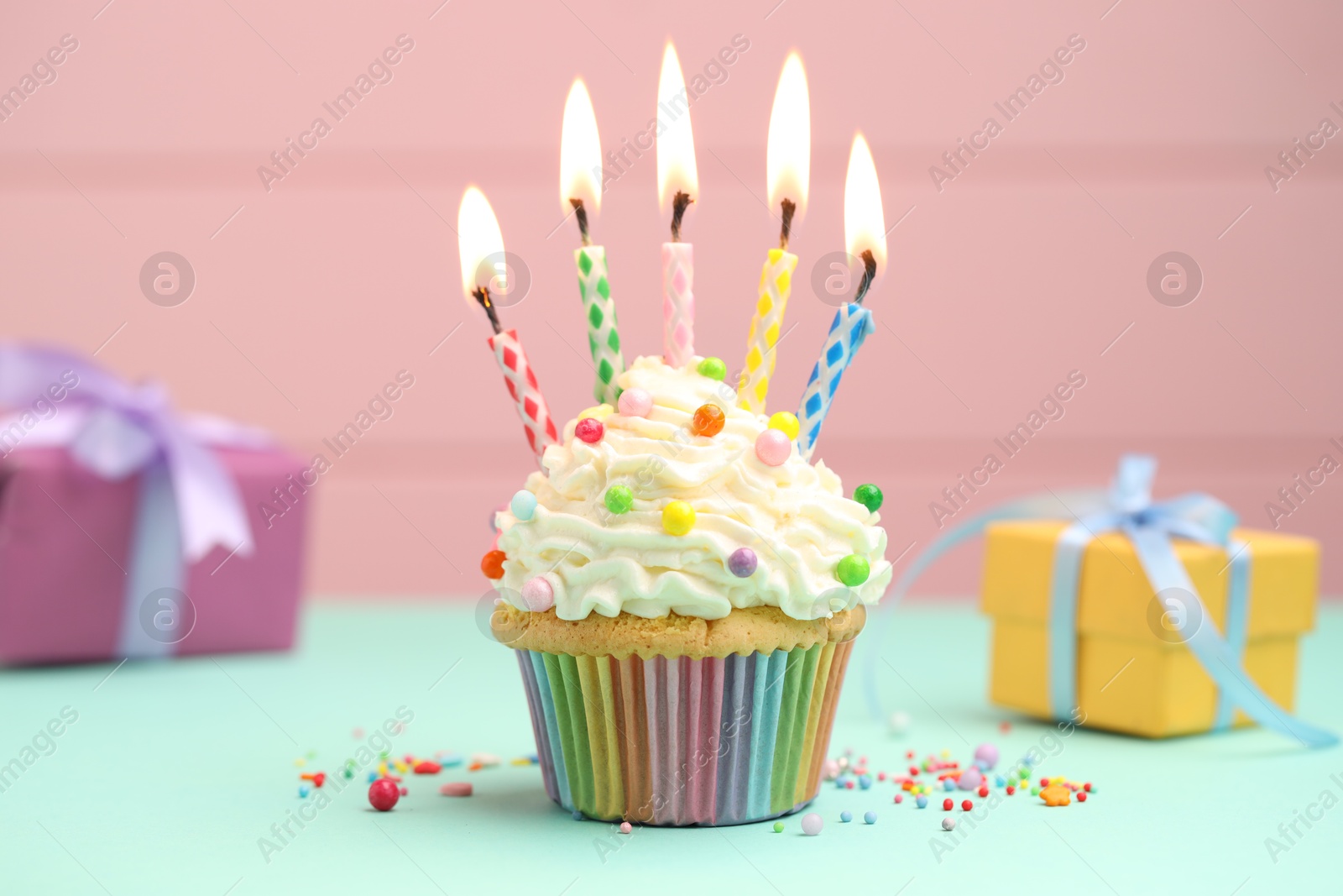 Photo of Burning candles in cupcake, sprinkles and gift boxes on turquoise table against pink background