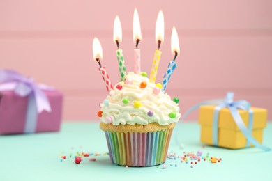 Photo of Burning candles in cupcake, sprinkles and gift boxes on turquoise table against pink background