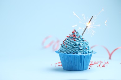 Photo of Burning sparkler in cupcake and sprinkles on light blue background, space for text