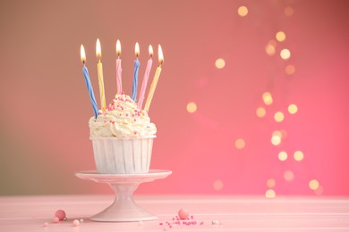 Photo of Burning candles in cupcake, sprinkles and party decor on table against color background with blurred lights, space for text