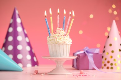 Photo of Burning candles in cupcake, sprinkles and party decor on pink table against color background with blurred lights