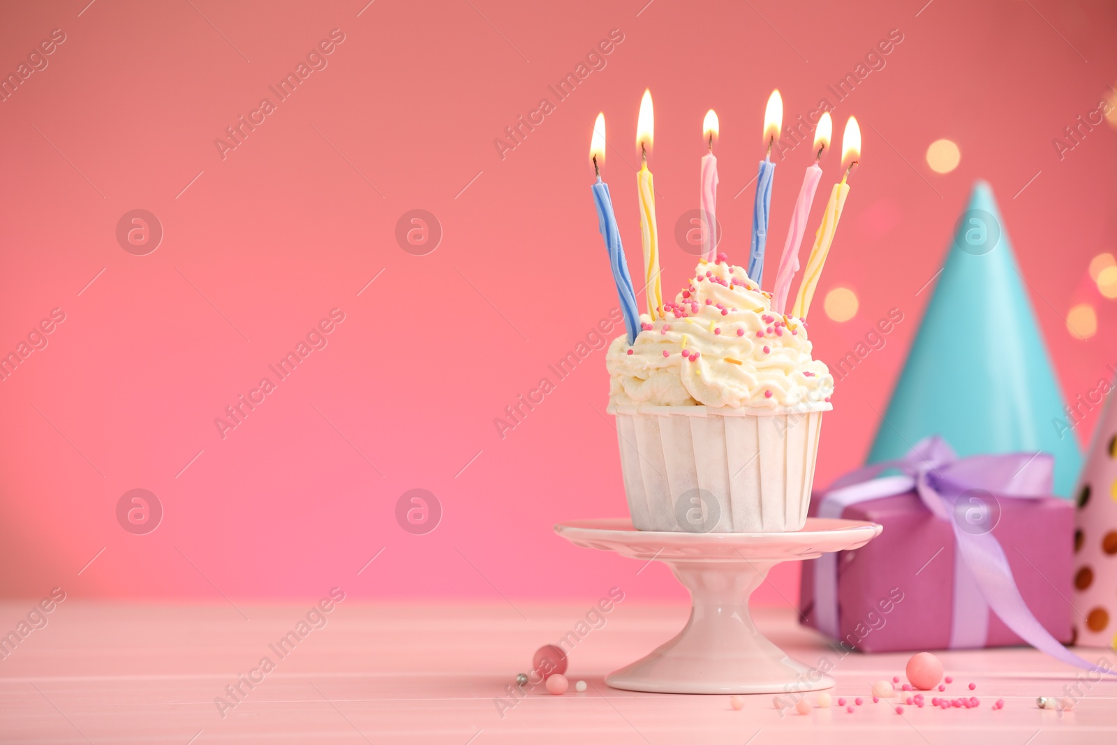 Photo of Burning candles in cupcake, sprinkles and party decor on pink table against color background with blurred lights, space for text