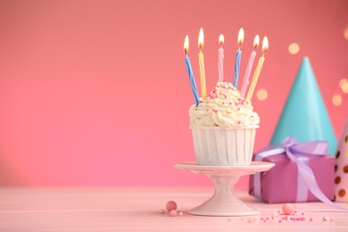 Photo of Burning candles in cupcake, sprinkles and party decor on pink table against color background with blurred lights, space for text