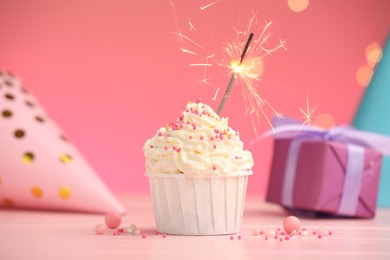 Photo of Burning sparkler in cupcake, sprinkles and party decor on pink table against color background with blurred lights