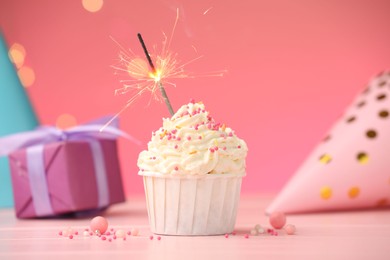 Photo of Burning sparkler in cupcake, sprinkles and party decor on pink table against color background with blurred lights