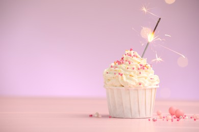 Photo of Burning sparkler in cupcake and sprinkles on pink table against violet background with blurred lights, space for text