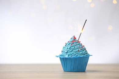 Photo of Burning sparkler in cupcake on wooden table against light background with blurred lights, space for text