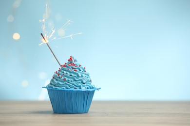 Photo of Burning sparkler in cupcake on wooden table against light blue background with blurred lights, space for text