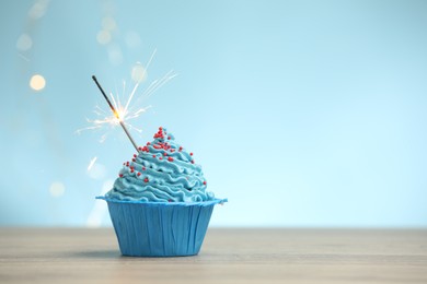 Photo of Burning sparkler in cupcake on wooden table against light blue background with blurred lights, space for text