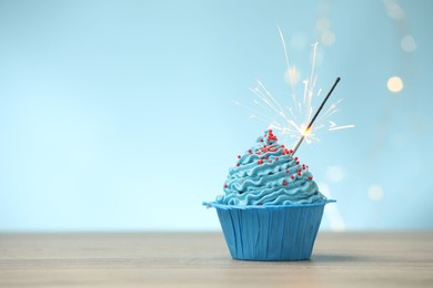 Photo of Burning sparkler in cupcake on wooden table against light blue background with blurred lights, space for text