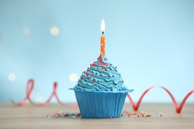 Photo of Burning candle in cupcake and sprinkles on wooden table against light blue background with blurred lights