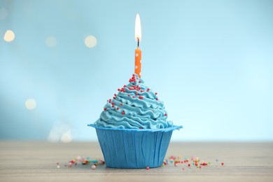 Photo of Burning candle in cupcake and sprinkles on wooden table against light blue background with blurred lights