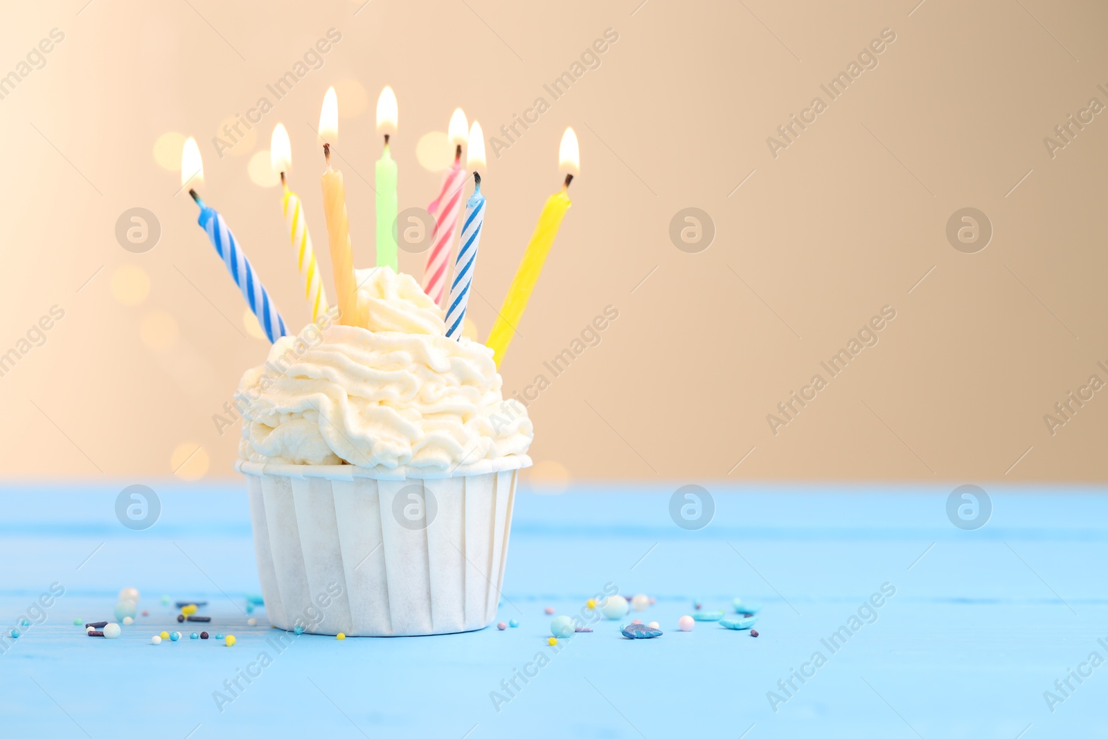 Burning candles in cupcake and sprinkles on light blue table against beige background with blurred lights Photo of Burning candles in cupcake and sprinkles on light blue table against beige background with blurred lights