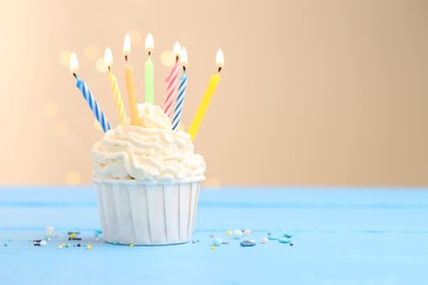 Photo of Burning candles in cupcake and sprinkles on light blue table against beige background with blurred lights