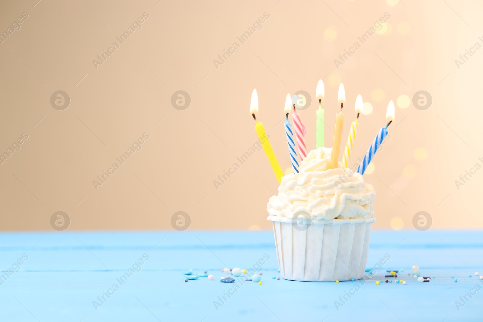 Burning candles in cupcake and sprinkles on light blue table against beige background with blurred lights Photo of Burning candles in cupcake and sprinkles on light blue table against beige background with blurred lights