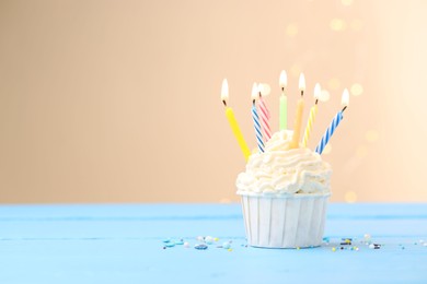 Photo of Burning candles in cupcake and sprinkles on light blue table against beige background with blurred lights