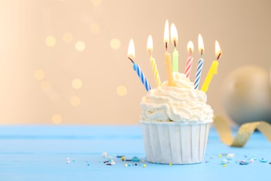 Photo of Burning candles in cupcake, sprinkles and party decor on light blue table against beige background with blurred lights, space for text