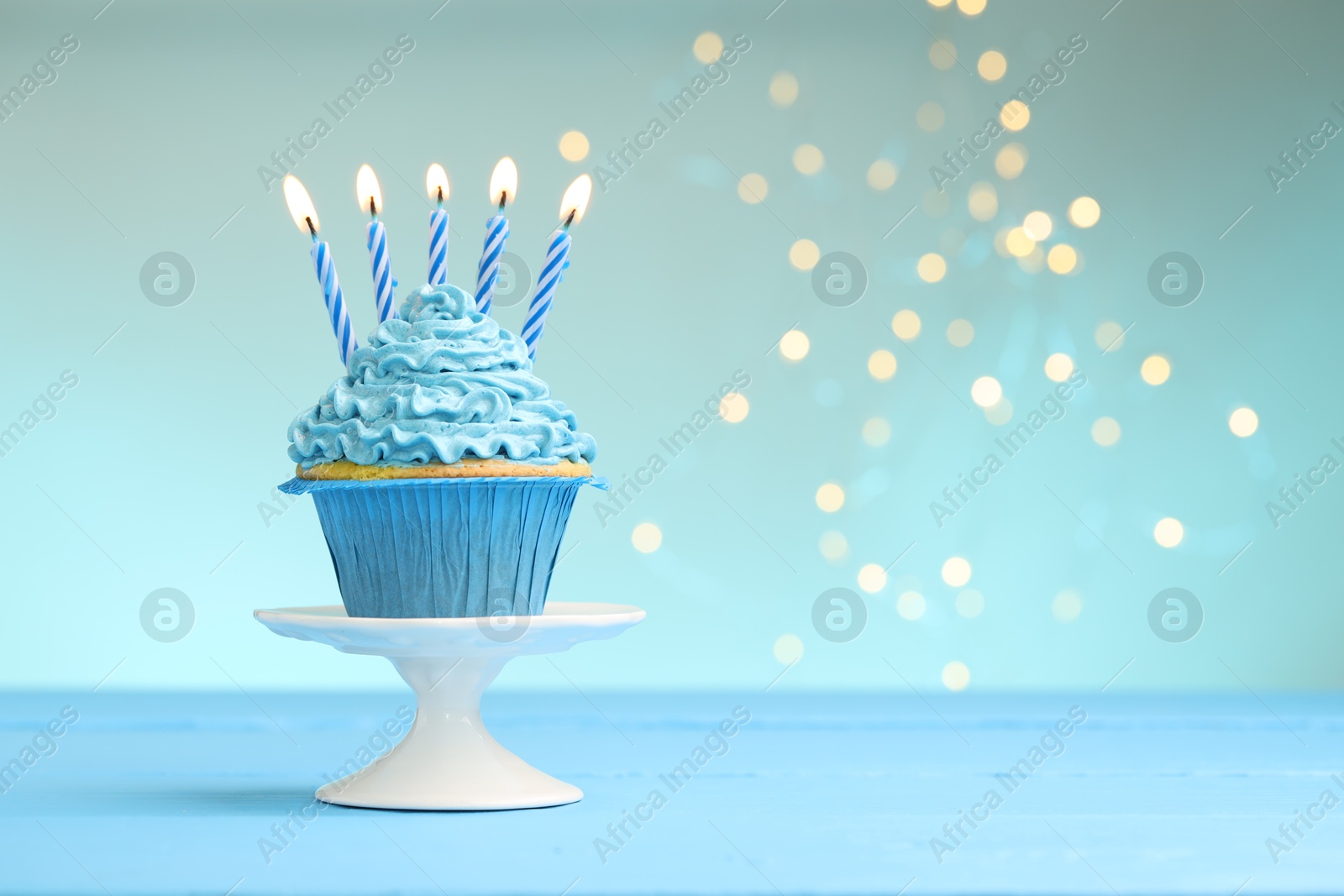 Photo of Burning candles in cupcake on light blue table against turquoise background with blurred lights, space for text