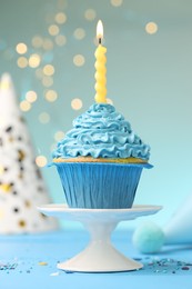 Photo of Burning candle in cupcake, sprinkles and party hats on light blue table against turquoise background with blurred lights