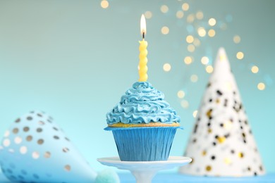 Photo of Burning candle in cupcake on cake stand and party hats against light blue background with blurred lights