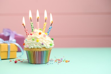 Photo of Burning candles in cupcake, sprinkles and gift boxes on turquoise table against pink background, space for text