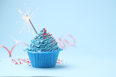 Photo of Burning sparkler in cupcake and sprinkles on light blue background, space for text