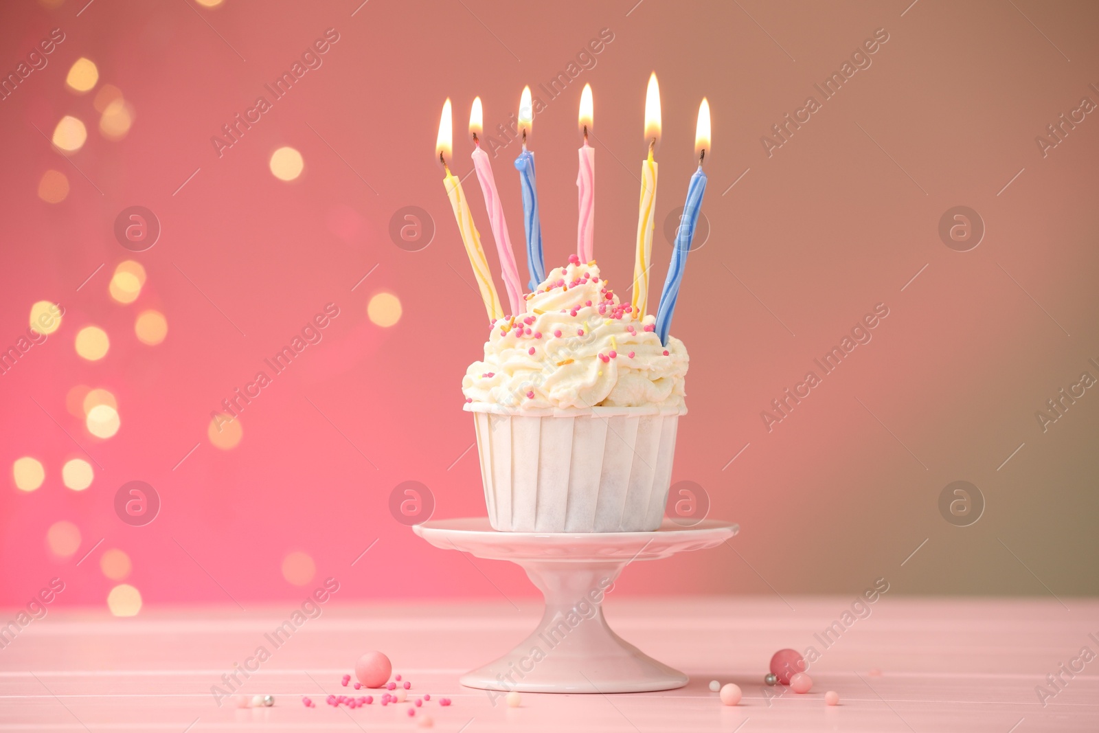 Photo of Burning candles in cupcake, sprinkles and party decor on table against color background with blurred lights