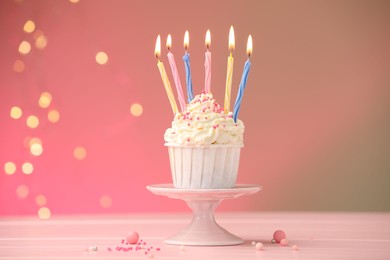 Photo of Burning candles in cupcake, sprinkles and party decor on table against color background with blurred lights