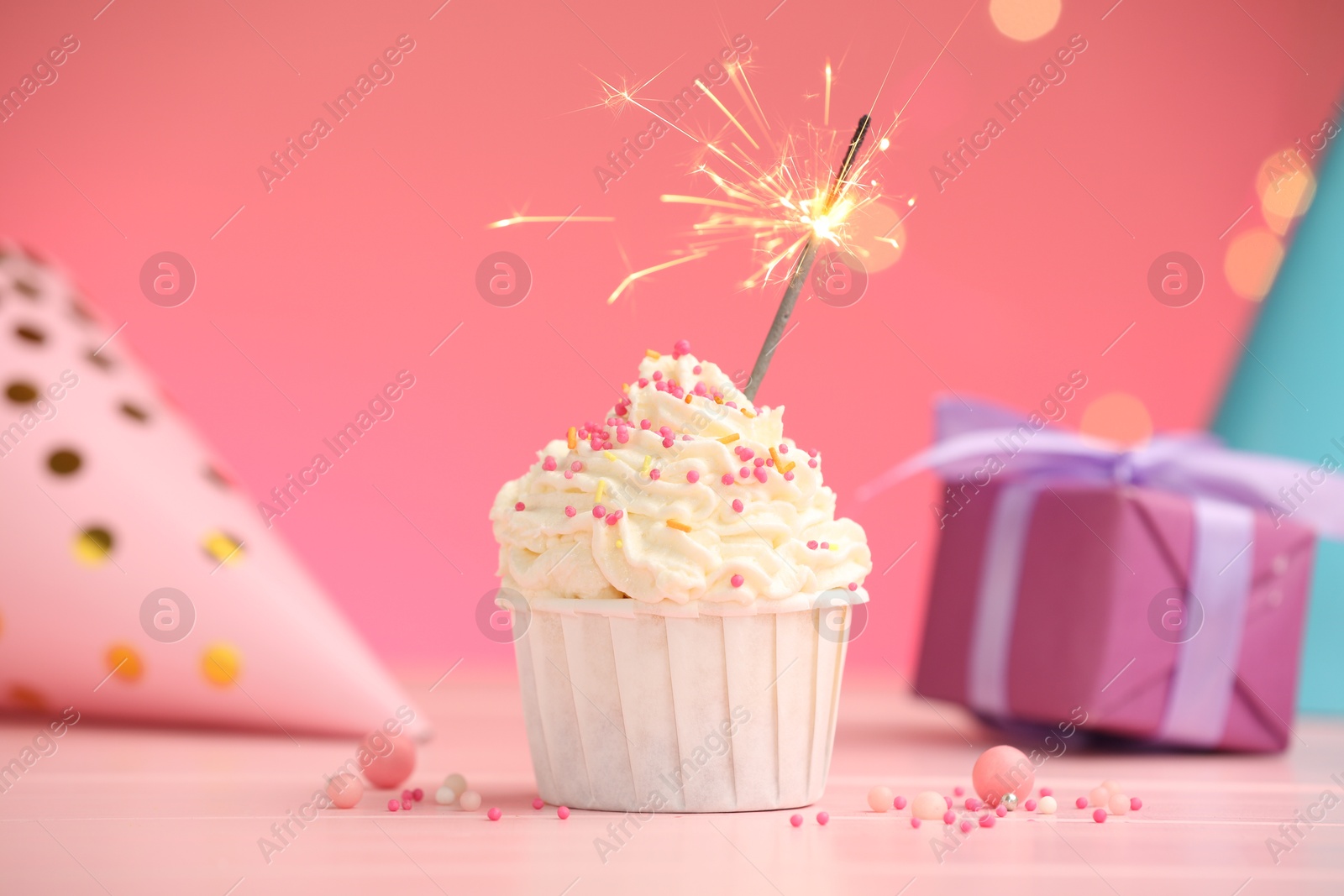 Photo of Burning sparkler in cupcake, sprinkles and party decor on pink table against color background with blurred lights